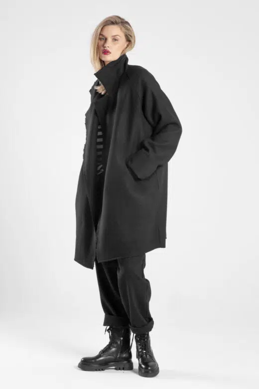 Modelka w czarnym płaszczu Niigata Black od delCane, ujęcie lekko bokiem z widoczną twarzą, ręka w kieszeni, kołnierz postawiony.