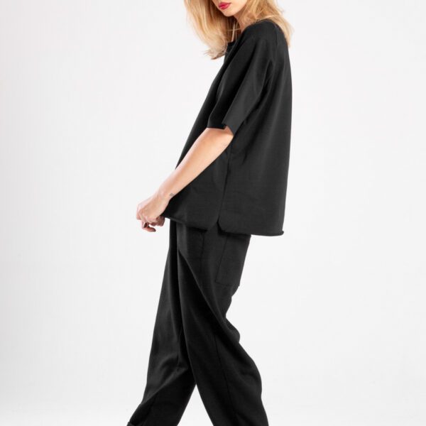 modelka w t-shircie Niigata Black, pełna sylwetka bokiem, patrzy w obiektyw, trzyma dół t-shirtu, widoczne boczne rozcięcia