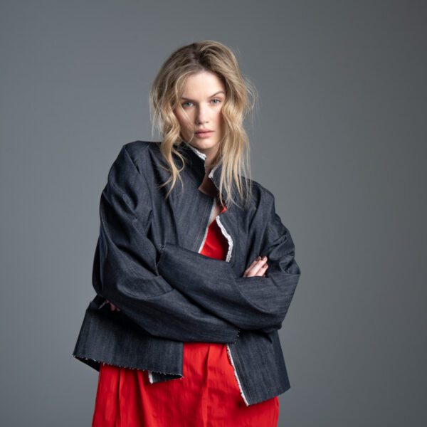 Modelka przodem prezentuje kurtkę Noda Jeans w ujęciu od kolan w górę, ręce ma splecione z przodu, w mocnym zbliżeniu wyraźnie widać kołnierz, detale z przodu oraz kolor jeansu.