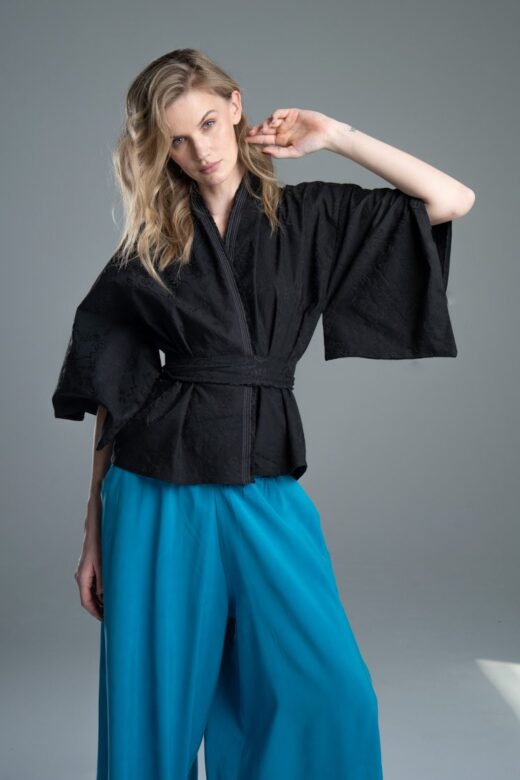 Modelka przodem prezentuje kimono Noda Black w ujęciu od kolan w górę, kimono jest związane, jedna ręka jest zgięta w łokciu i uniesiona, wyraźnie widać całe kimono oraz detal rękawa.