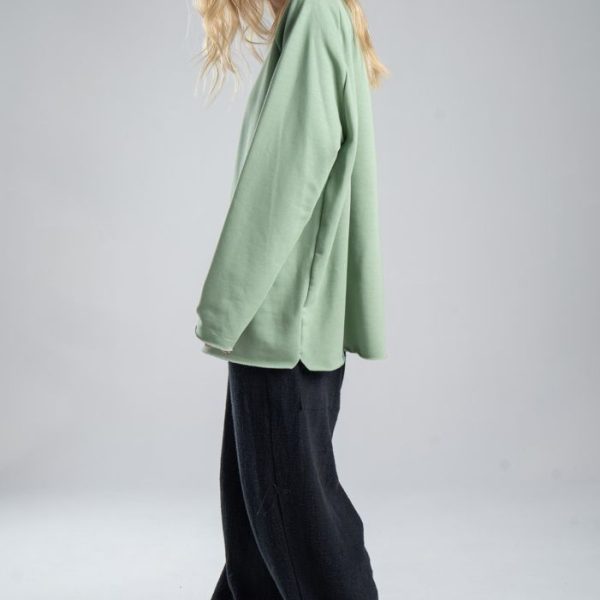 Bluza Mino Green delCane w miętowym odcieniu zieleni. Modelka bokiem, w pełnej sylwetce, patrzy w obiektyw. Wyraźnie widoczny bok bluzy.