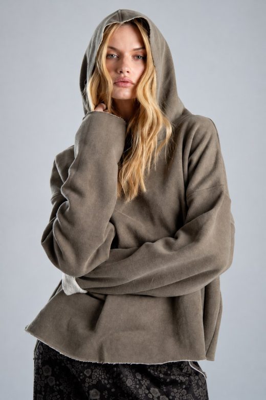 Bluza Mino Stone delCane w szarobeżowym kolorze. Zbliżenie na przód bluzy i kaptur założony na głowę. Modelka patrzy w obiektyw, z jedną dłonią uniesioną przy twarzy.