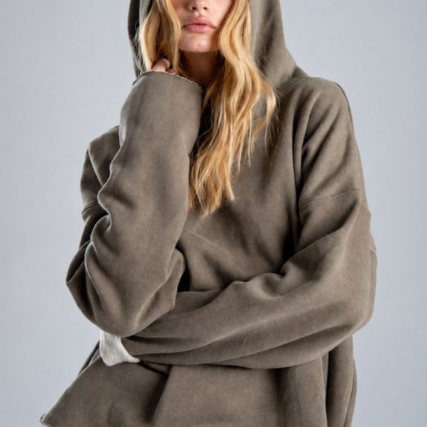 Bluza Mino Stone delCane w szarobeżowym kolorze. Zbliżenie na przód bluzy i kaptur założony na głowę. Modelka patrzy w obiektyw, z jedną dłonią uniesioną przy twarzy.