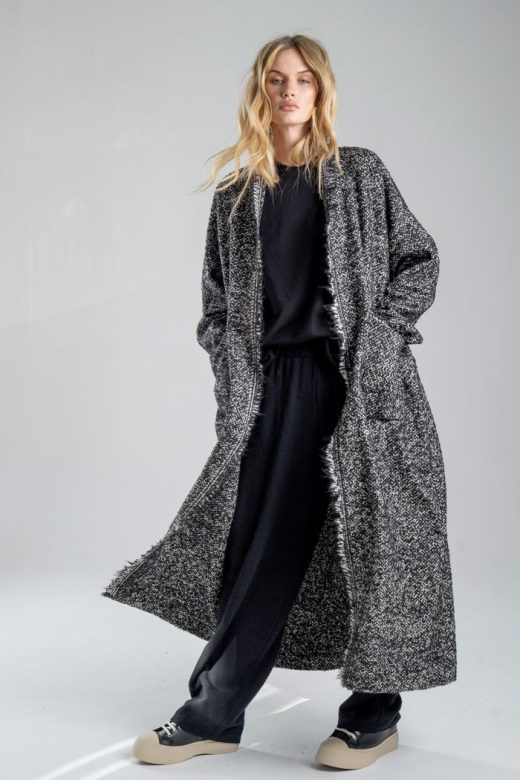 Cardigan Mino Wool delCane w czarno-białym melanżu. Modelka przodem, w pełnej sylwetce, w ruchu, z rękami w kieszeniach cardiganu. Wyraźnie widoczny cały przód.