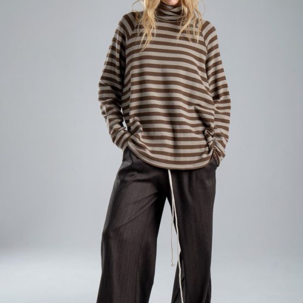 Sweter z golfem Mino Stripes delCane w beżowo-brązowe paski. Modelka w pełnej sylwetce, ustawiona przodem, z rękami w kieszeniach spodni, patrzy w obiektyw.