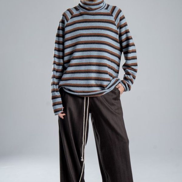 Sweter z golfem Mino Wool delCane w niebiesko-brązowe paski. Modelka w pełnej sylwetce, ustawiona przodem, z jedną ręką w kieszeni spodni i wzrokiem skierowanym w bok.