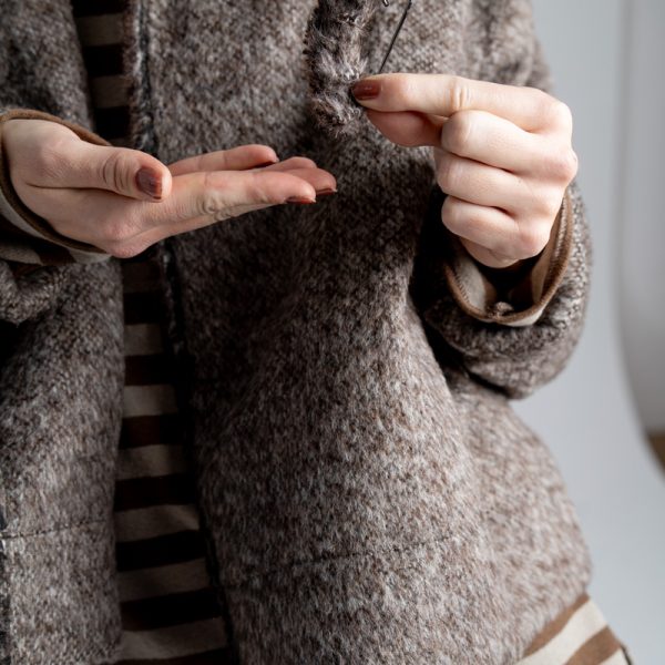 Kurtka Mino Wool delCane w melanżowy wzór w odcieniach brązu. Zbliżenie na detal z obszytą wełną agrafką trzymaną w dłoniach modelki.