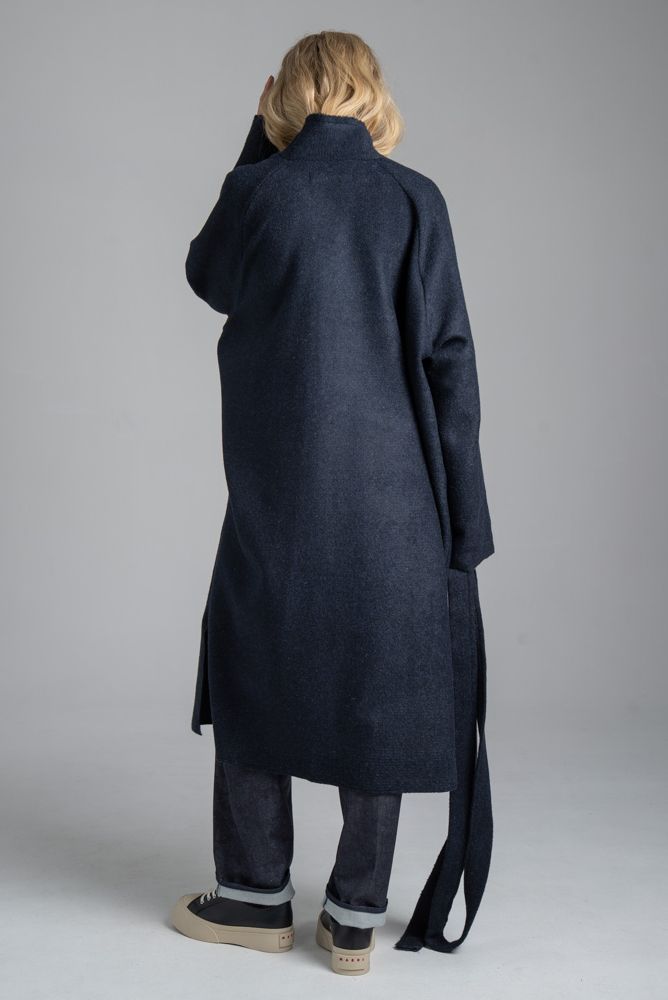 Granatowy płaszcz Mino Wool delCane, widok od tyłu. Modelka w pełnej sylwetce, w swobodnej pozie, trzyma w jednej ręce rozwiązany pasek płaszcza.